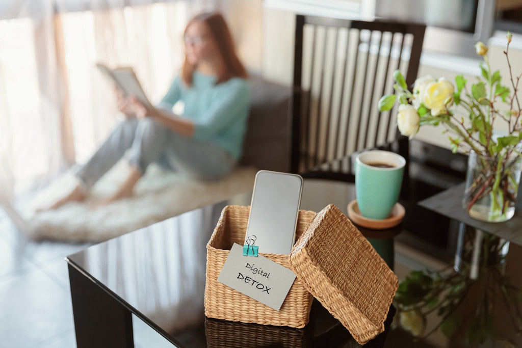 Smartphone liegt in einem geflochtenen Korb auf einem Tisch; daneben steht eine Kaffeetasse und eine Vase mit Blumen. Im unscharfen Hintergrund liest eine Frau ein Buch. Das Bild symbolisiert Digital Detox.