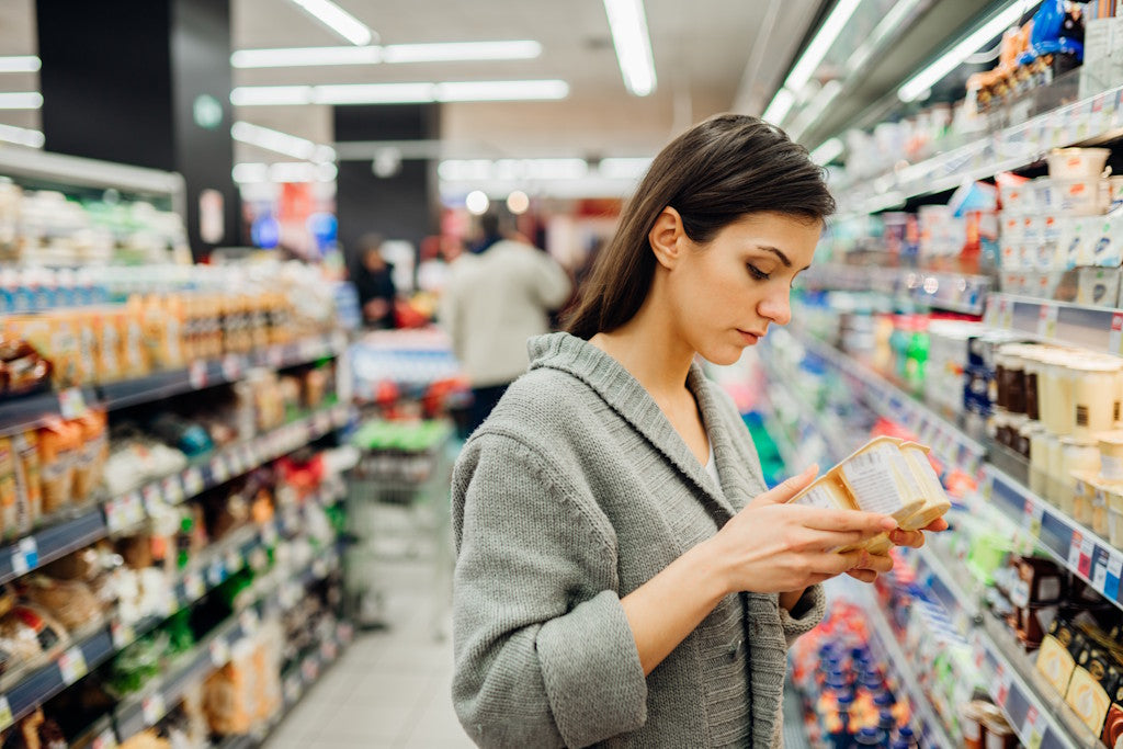Frau steht vor einem Supermarktregal und liest sich die Angaben auf einer Joghurt-Packung durch
